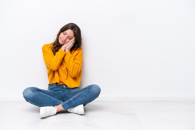 Young caucasian woman sitting on the floor isolated on white wall making sleep gesture in dorable expression