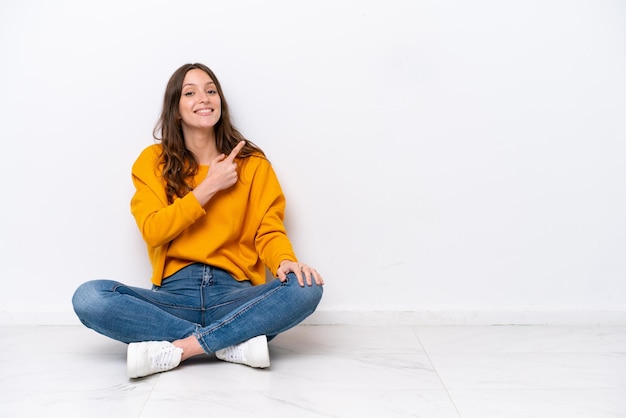 Young caucasian woman sitting on the floor isolated on white wall pointing to the side to present a product