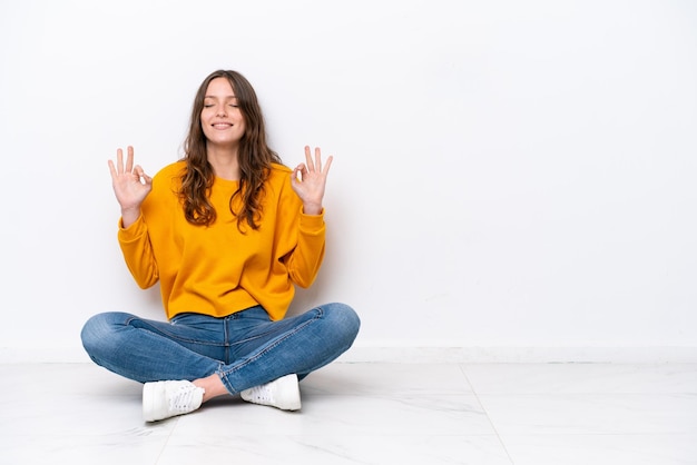 Young caucasian woman sitting on the floor isolated on white wall in zen pose