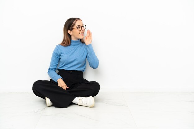 Young Caucasian woman sitting on the floor shouting with mouth wide open to the lateral