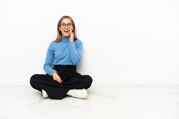 Young Caucasian woman sitting on the floor shouting with mouth wide open