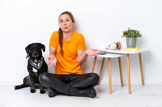 Young caucasian woman sitting on the floor with his puppy isolated on white background making doubts gesture while lifting the shoulders