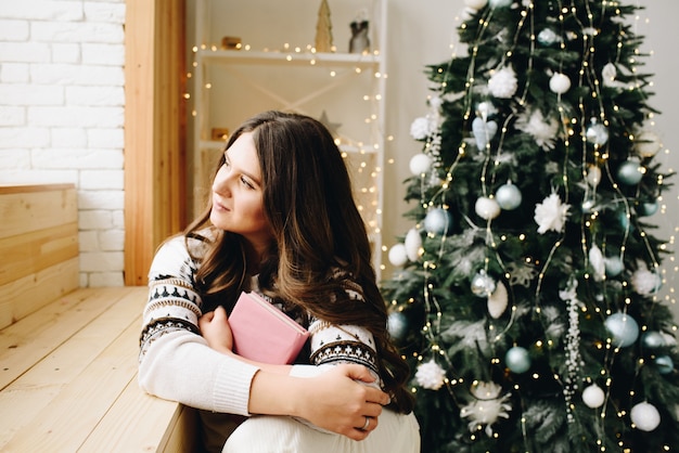 Young Caucasian woman sitting next to lovely decorated Christmas Tree holding a book and dreaming