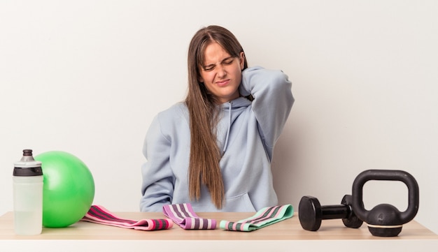 Young caucasian woman sitting at a table with sport equipment isolated on white background suffering neck pain due to sedentary lifestyle.