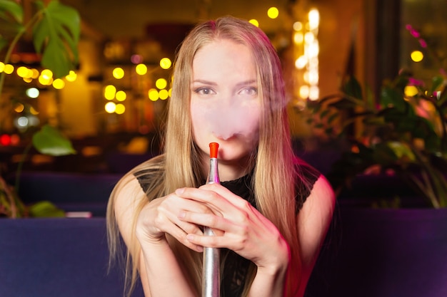 Young caucasian woman smokes a hookah or shisha in the night club or bar smoke