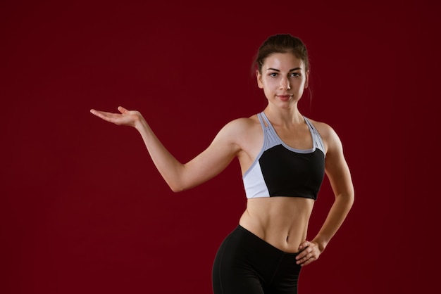 Young caucasian woman in sportswear on a red background shows her hand to the side concept for adver...