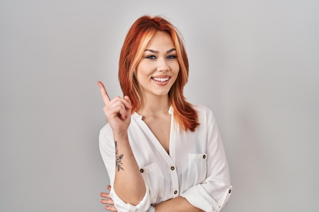 Young caucasian woman standing over isolated background with a big smile on face, pointing with hand finger to the side looking at the camera.