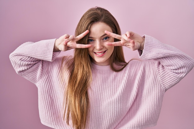 Young caucasian woman standing over pink background doing peace symbol with fingers over face smiling cheerful showing victory