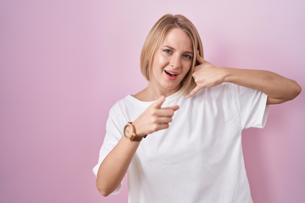 Young caucasian woman standing over pink background smiling doing talking on the telephone gesture and pointing to you. call me.