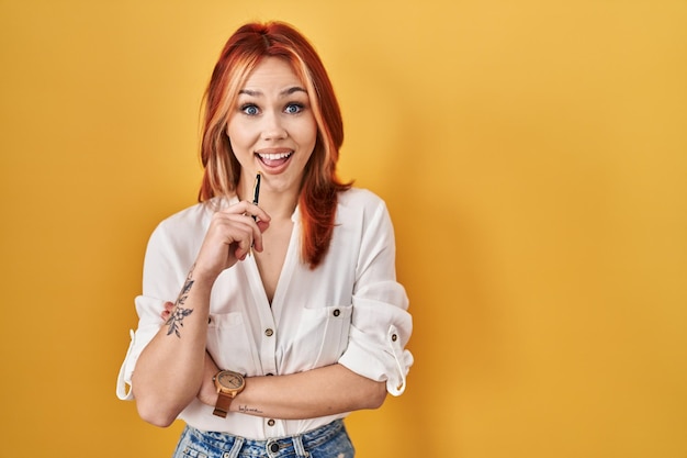 Young caucasian woman standing over yellow background holding pen celebrating crazy and amazed for success with open eyes screaming excited