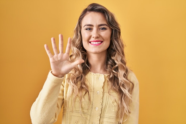 Young caucasian woman standing over yellow background showing and pointing up with fingers number five while smiling confident and happy