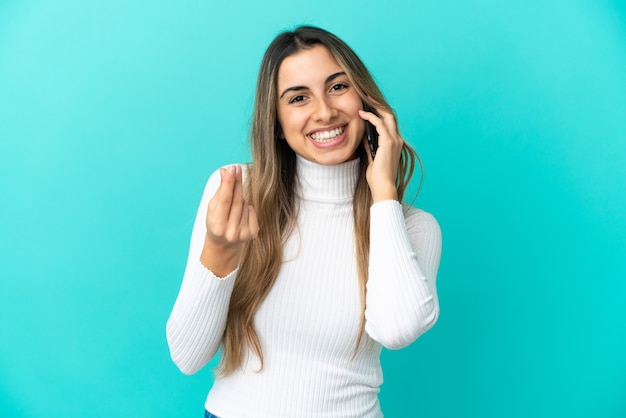 Young caucasian woman using mobile phone isolated on blue background making money gesture