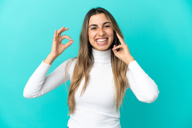 Young caucasian woman using mobile phone isolated on blue background showing ok sign with fingers