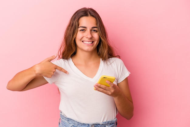 Young caucasian woman using mobile phone isolated on pink background  person pointing by hand to a shirt copy space, proud and confident