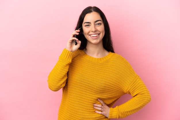 Young caucasian woman using mobile phone isolated on pink background posing with arms at hip and smiling