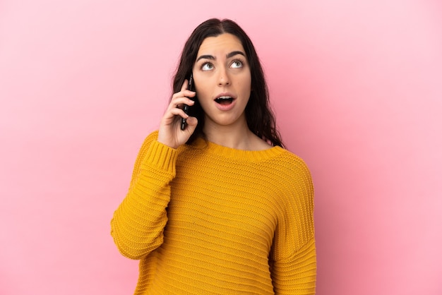 Young caucasian woman using mobile phone isolated on pink wall looking up and with surprised expression