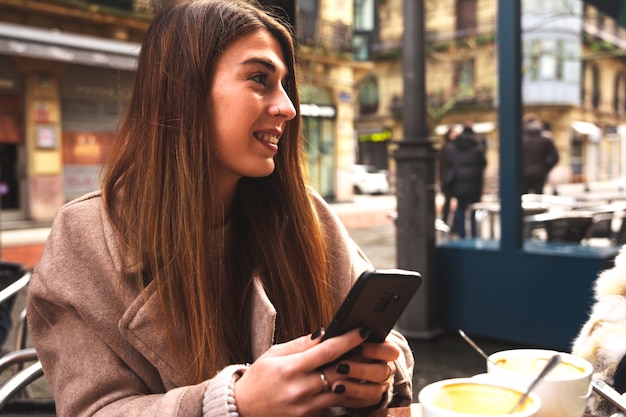 Young caucasian woman using a mobile phone while having breakfast at a terrace on a winter morning