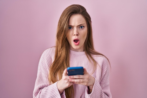 Young caucasian woman using smartphone typing message in shock face, looking skeptical and sarcastic, surprised with open mouth