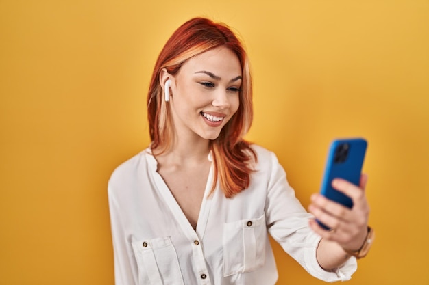 Young caucasian woman using smartphone wearing earphones looking positive and happy standing and smiling with a confident smile showing teeth