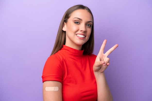 Young caucasian woman wearing band aid isolated on purple background smiling and showing victory sign