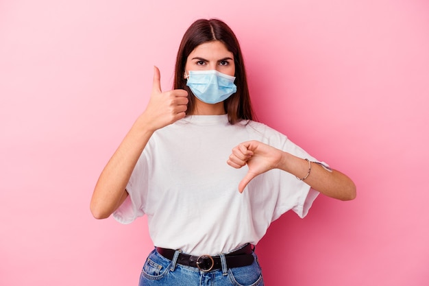 Young caucasian woman wearing a mask for virus isolated on pink wall showing thumbs up and thumbs down, difficult choose concept