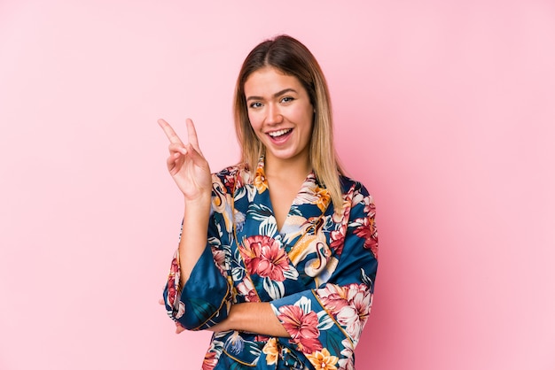 Young caucasian woman wearing pajamas joyful and carefree showing a peace symbol with fingers.