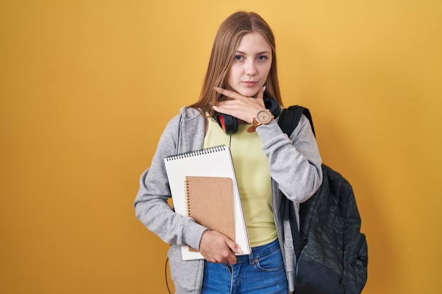 Young caucasian woman wearing student backpack and holding books cutting throat with hand as knife threaten aggression with furious violence