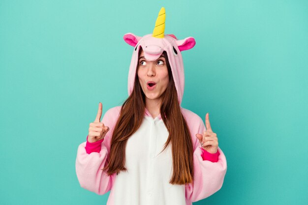 Young caucasian woman wearing a unicorn pajama isolated on blue background pointing upside with opened mouth.