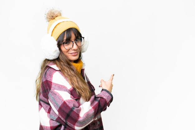 Young caucasian woman wearing winter muffs over isolated white background pointing back