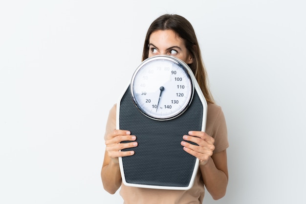 Young caucasian woman on white wall with weighing machine and hiding behind it