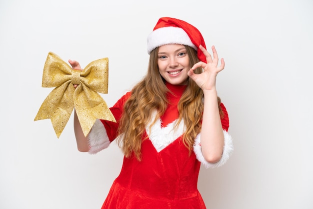 Young caucasian woman with Christmas dress holding Christmas bow isolated on white background showing ok sign with fingers