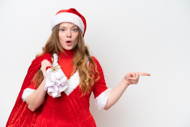 Young caucasian woman with Christmas dress holding Christmas sack isolated on white background surprised and pointing side