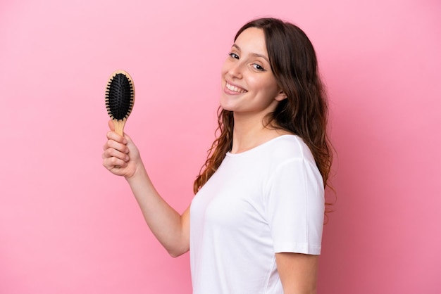 Photo young caucasian woman with hair comb isolated on pink background smiling a lot