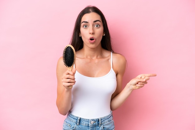 Young caucasian woman with hair comb isolated on pink background surprised and pointing side