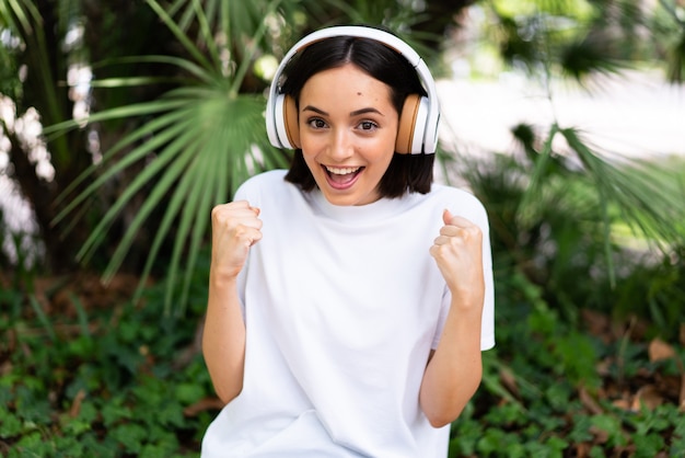 Young caucasian woman with headphones at outdoors celebrating a victory in winner position