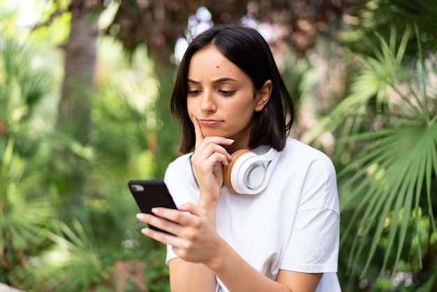 Young caucasian woman with headphones at outdoors thinking and sending a message