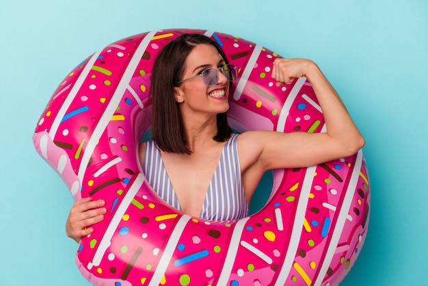 Young caucasian woman with inflatable donut isolated on blue background raising fist after a victory, winner concept.