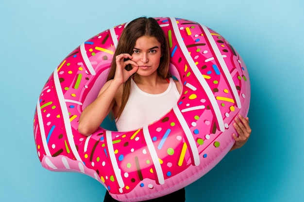 Young caucasian woman with inflatable donut isolated on blue background with fingers on lips keeping a secret.