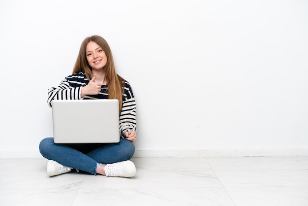 Young caucasian woman with a laptop sitting on the floor isolated on white background giving a thumbs up gesture