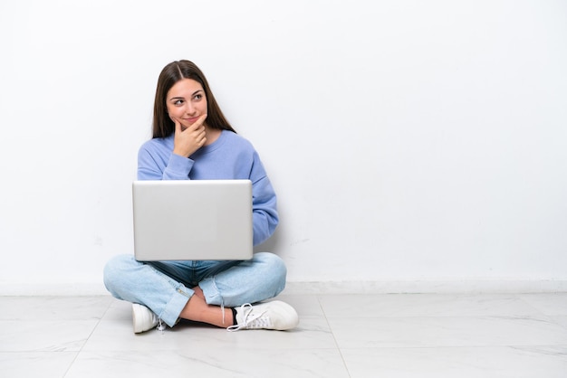 Young caucasian woman with laptop sitting on the floor isolated on white background having doubts and with confuse face expression