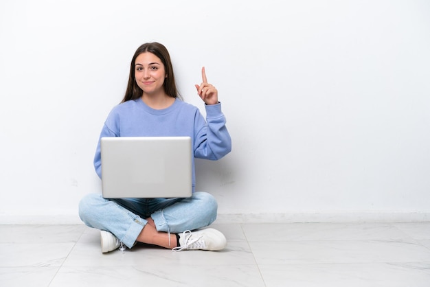 Young caucasian woman with laptop sitting on the floor isolated on white background pointing with the index finger a great idea