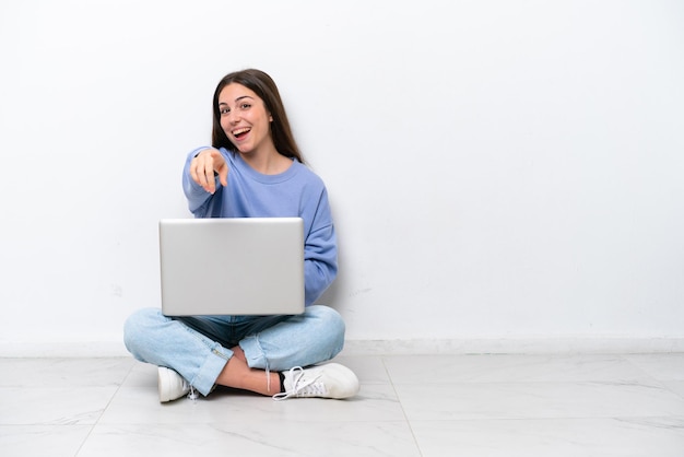 Young caucasian woman with laptop sitting on the floor isolated on white background points finger at you with a confident expression