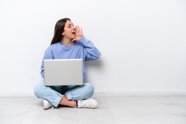 Young caucasian woman with laptop sitting on the floor isolated on white background shouting with mouth wide open to the lateral