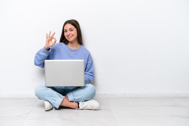 Young caucasian woman with laptop sitting on the floor isolated on white background showing ok sign with fingers