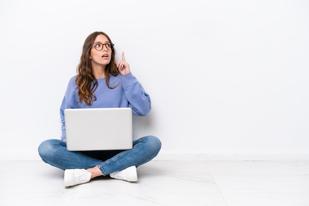 Young caucasian woman with a laptop sitting on the floor isolated on white background thinking an idea pointing the finger up