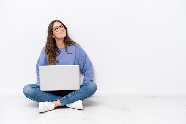 Young caucasian woman with a laptop sitting on the floor isolated on white background thinking an idea while looking up