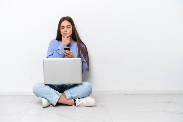 Young caucasian woman with laptop sitting on the floor isolated on white background thinking and sending a message