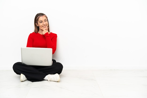 Young caucasian woman with a laptop sitting on the floor looking to the side