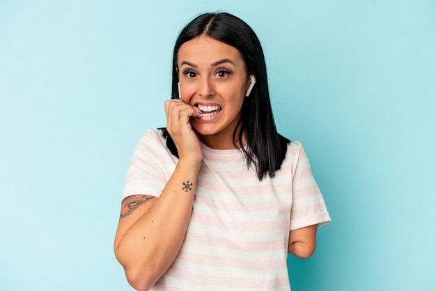 Young caucasian woman with one arm listening to music with wireless headphones isolated on blue background biting fingernails, nervous and very anxious.