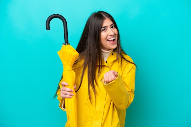 Young caucasian woman with rainproof coat and umbrella isolated on blue background celebrating a victory